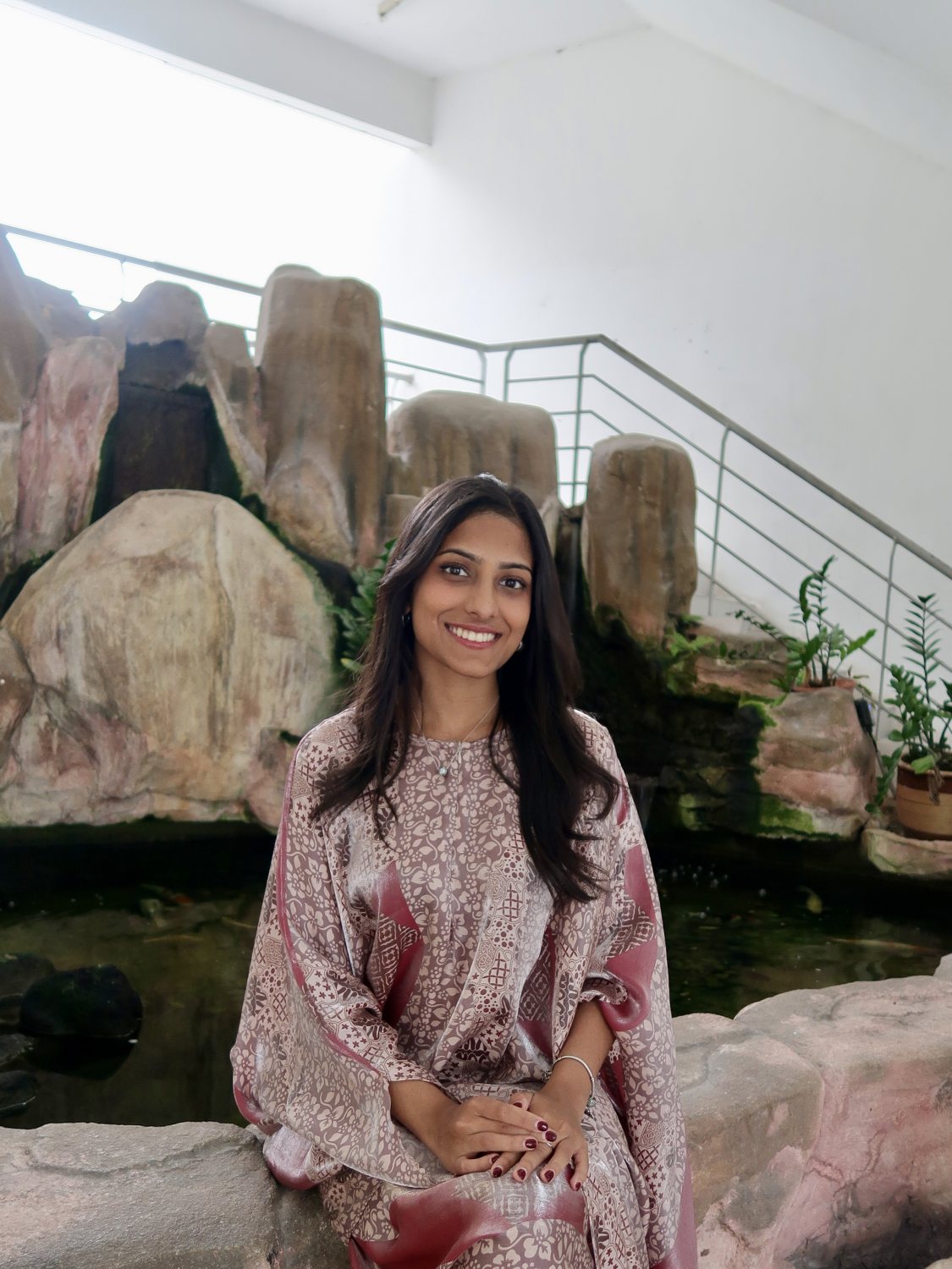 Isheta Shah, smiling, seated in front of a rock water feature